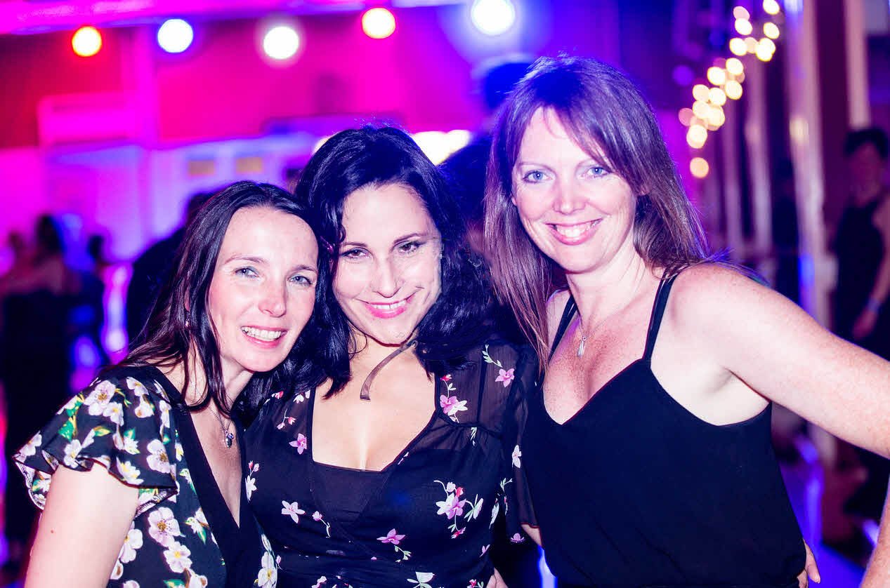Three smiling women posing together at a Jive Addiction event, with colorful lights in the background.
